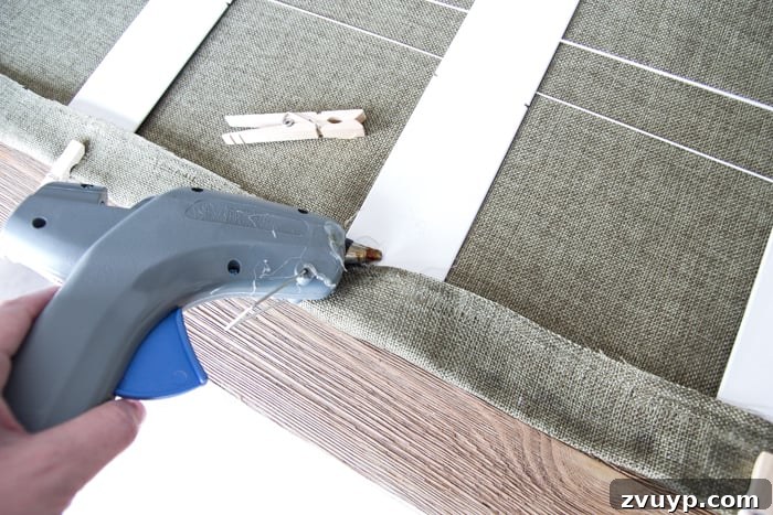 Burlap Roman Shades A Clever Blind Transformation 12 Hot glue being applied to the edge of a slat, with fabric pressed over it and secured by clothespins, demonstrating the crucial step of attaching fabric to slats for DIY Roman shades.