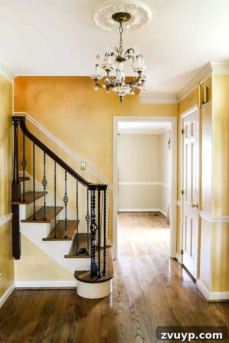 A Fresh Welcome: Our Neutral Foyer Makeover 2 Dated yellow foyer with faux aging finish before renovation. Shows high ceilings, staircase, and original almond trim.