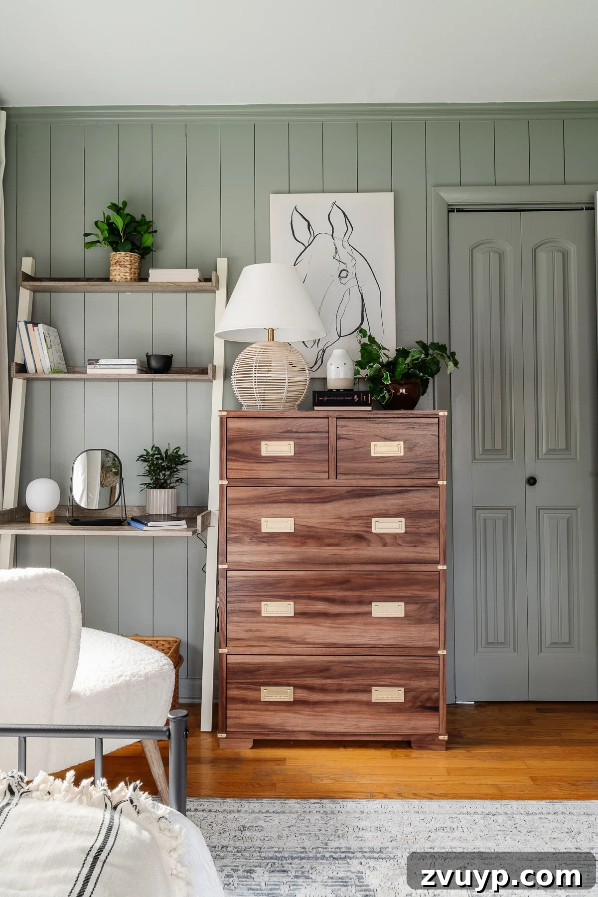 A serene sage green bedroom with matching walls and trim, featuring a wooden dresser, a leaning desk, and a white boucle desk chair, all contributing to an expansive atmosphere.