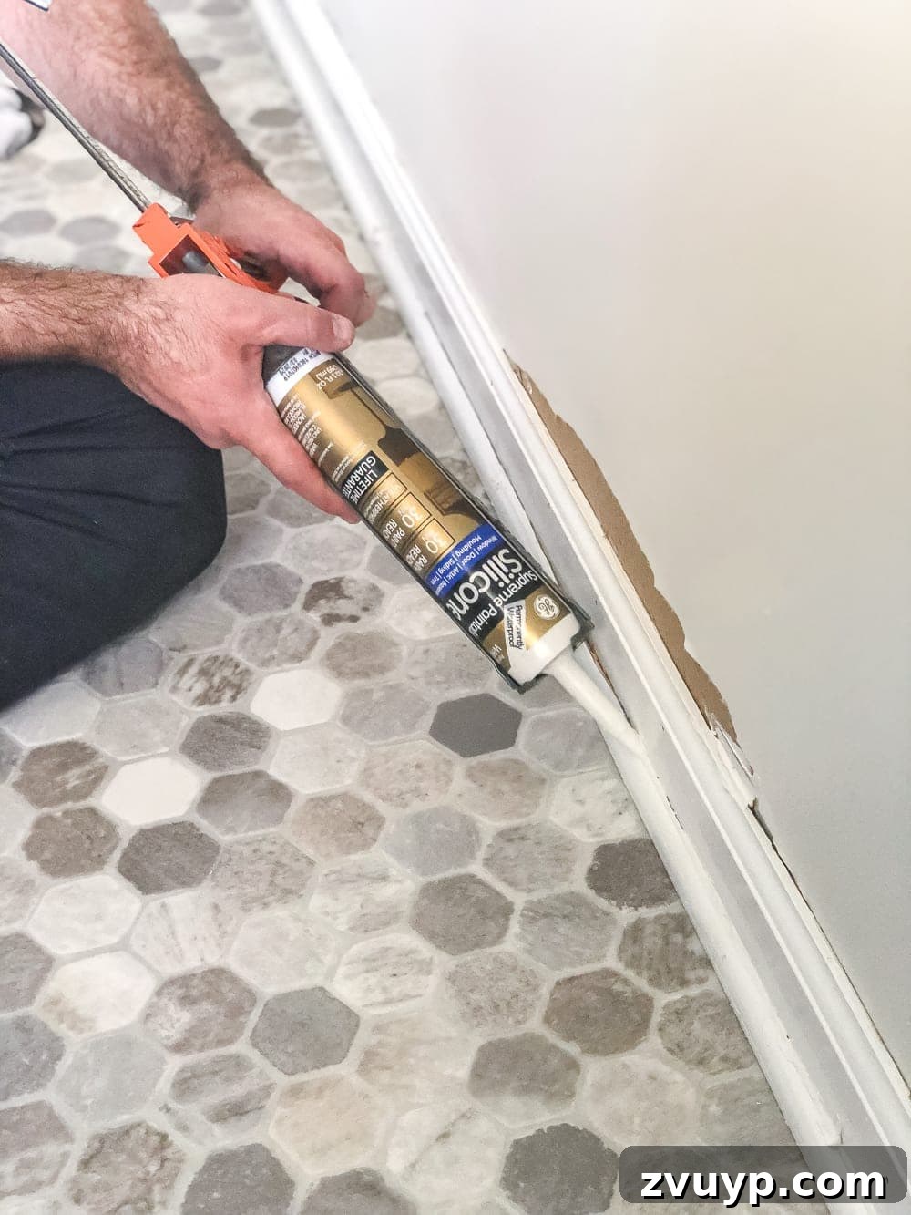 DIY Floor Makeover Seamless Sheet Vinyl Over Tile 17 A person applying white silicone caulk neatly along the joint where the newly reinstalled baseboard meets the wall.