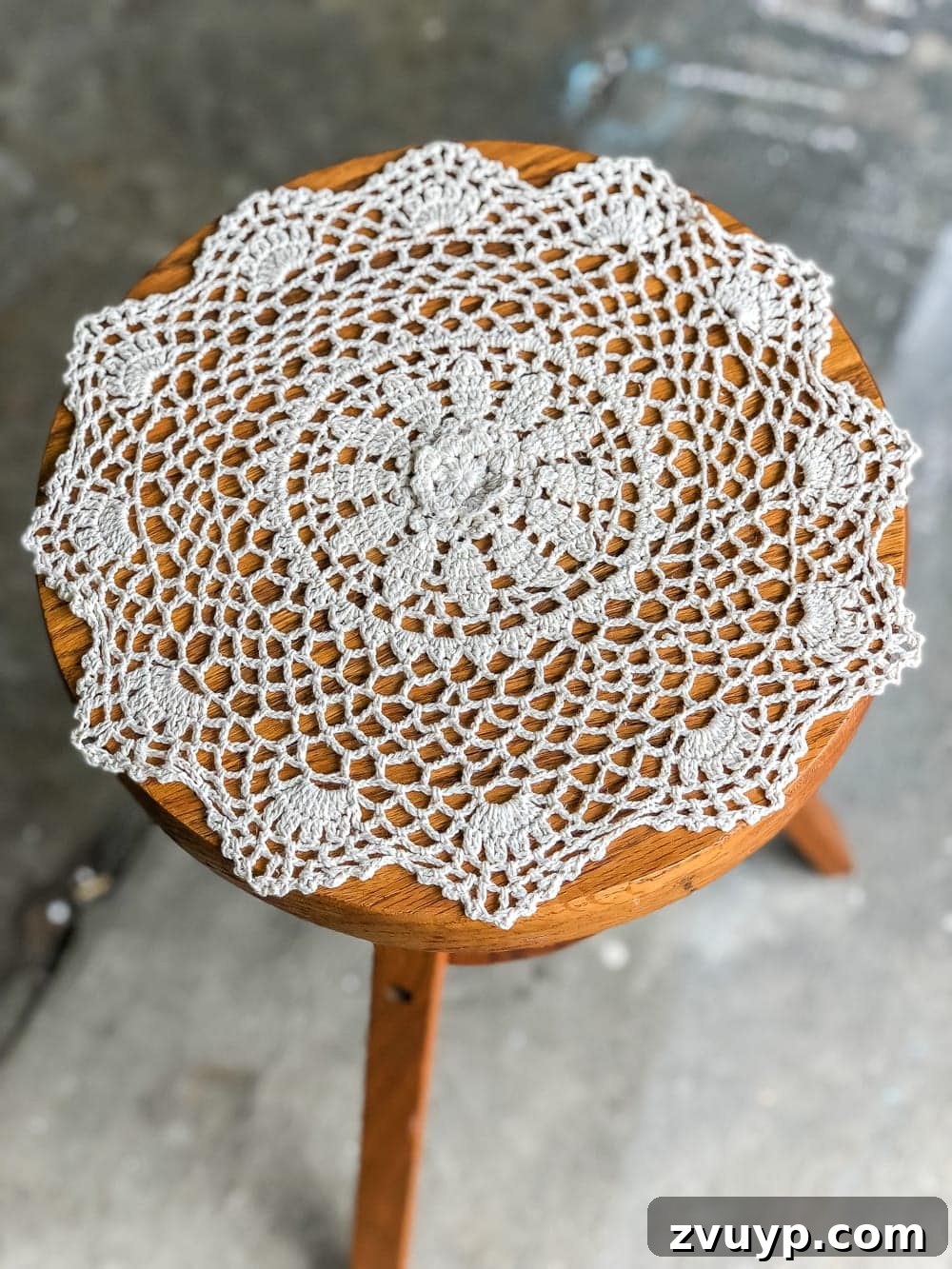 Preparing the thrifted piano stool for a DIY makeover. The stool top is partially taped off, revealing a natural wood section next to the area designated for doily stenciling and white spray paint.