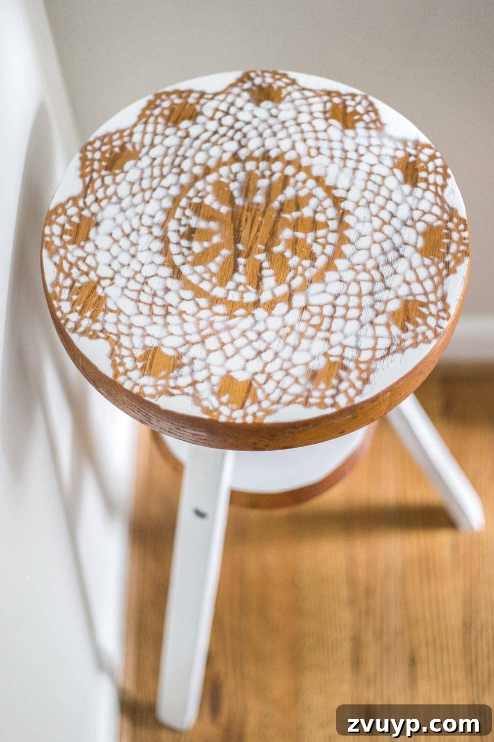 A charming DIY plant stand, repurposed from a thrifted piano stool with a doily stencil and white paint, elegantly styled with a realistic faux plant in a dimly lit hallway corner.