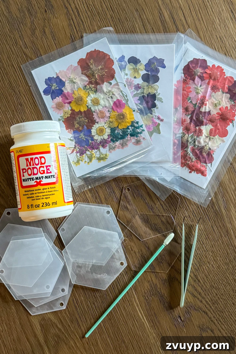 Surprisingly Simple Pressed Flower Ornaments 4 All the necessary craft supplies laid out: clear acrylic tags, a bottle of matte Mod Podge, a craft brush, a roll of thin ribbon, and a variety of colorful pressed flowers, ready for making DIY Christmas ornaments.